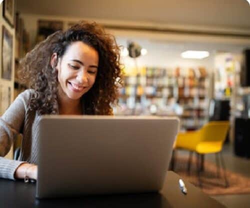 A student studies in the library, smiling as they look at their laptop.